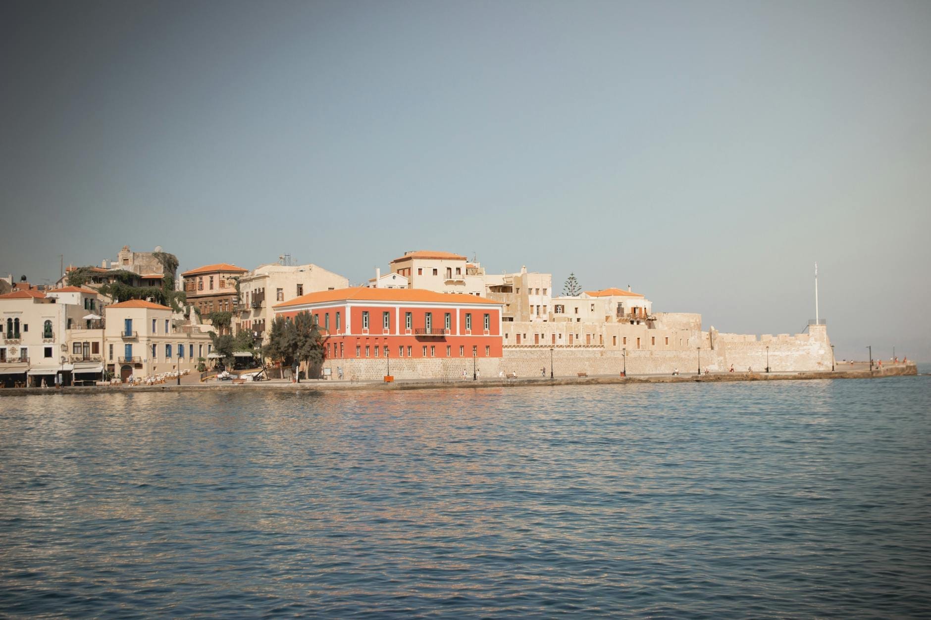 Vista del porto veneziano di Chania con la Fortezza Firka e l'edificio del Museo Marittimo, riflessi nell'acqua calma sotto un cielo tenue.