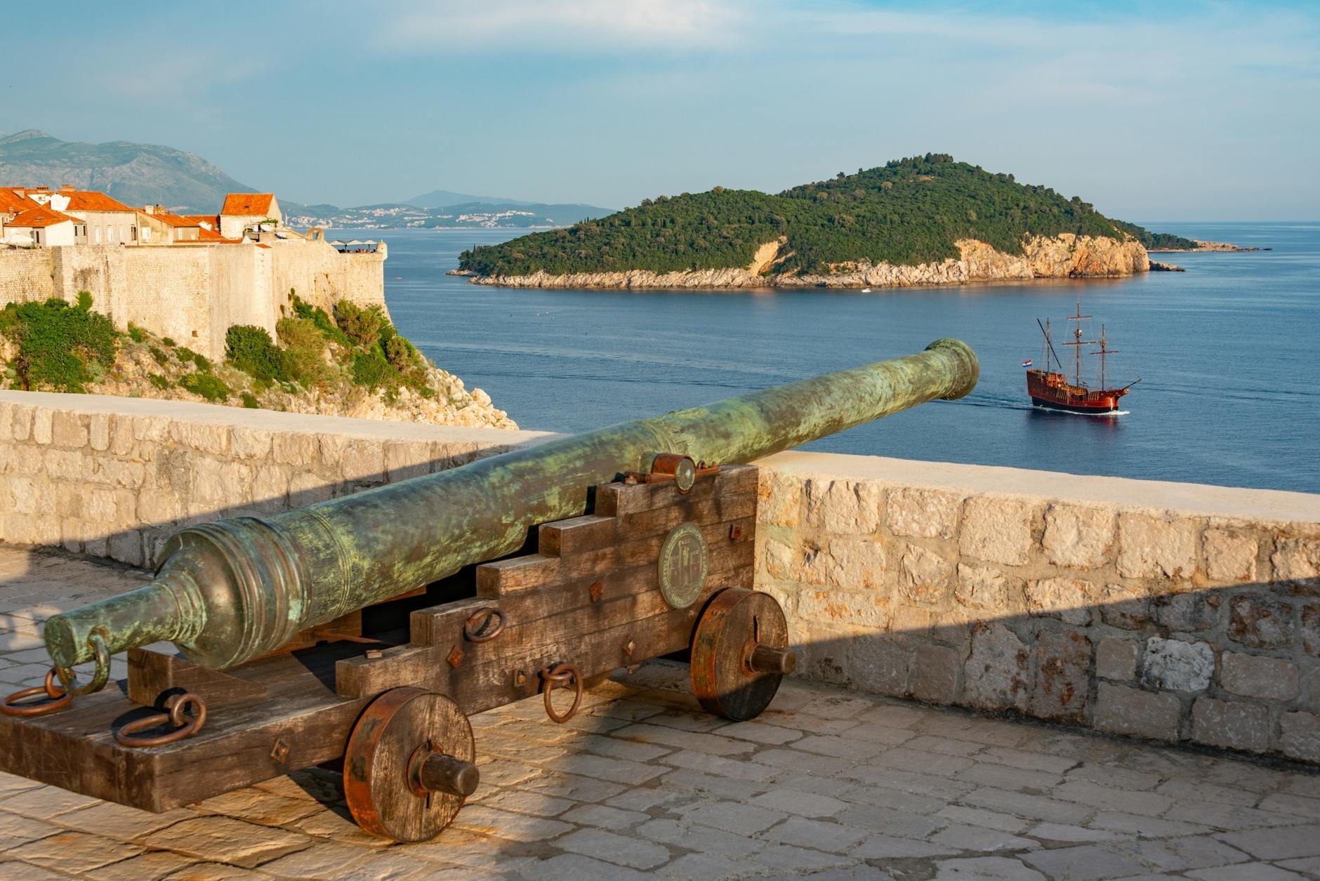 Antiguo cañón de bronce sobre la muralla de piedra de la fortaleza de Dubrovnik, con vistas al azul del Adriático, las fortificaciones, una isla verde y un velero a lo lejos.