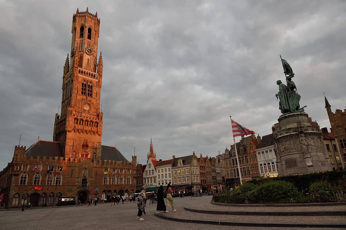 Wide view of Bruges’ Markt with the 13th-century Belfry and monumental statue, surrounded by medieval guild houses and bustling square at sunset.