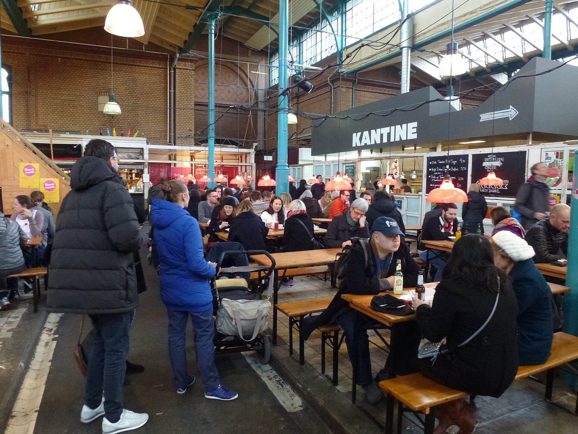 I visitatori si riuniscono attorno a lunghi tavoli in legno all'interno di Markthalle Neun, con soffitti alti, mattoni a vista e vivaci bancarelle gastronomiche sullo sfondo.