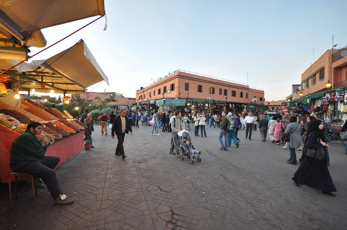 มุมมองกว้างของจัตุรัสตลาดเมดินาในเมือง Marrakech ช่วงกลางวัน พร้อมแผงขายอาหารและสินค้า และผู้คนที่เดินและช้อปปิ้ง
