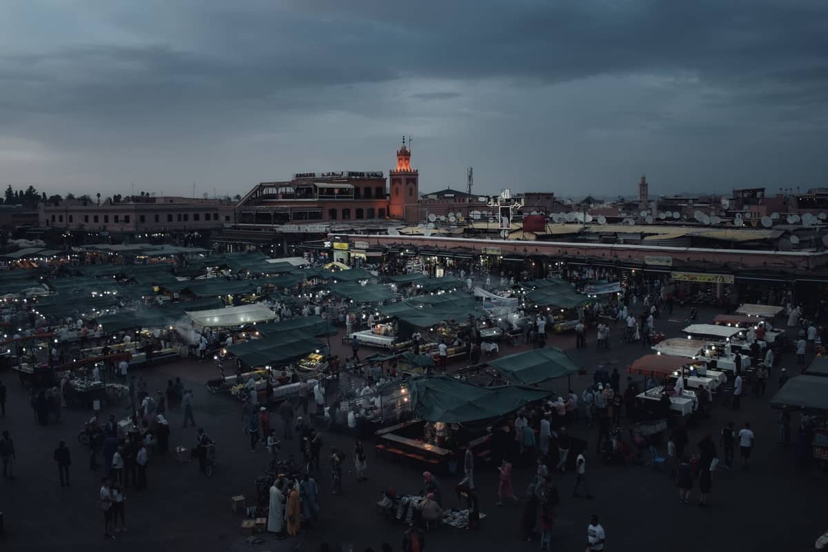 ภาพถ่ายทางอากาศยามเย็นของจัตุรัสหลักในเมือง Marrakech พร้อมแผงขายอาหารในตลาดหลายสิบแผงที่สว่างไสวขึ้นขณะที่ฝูงชนมารวมตัวกันในยามพลบค่ำ