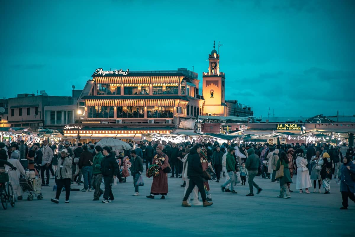 ฝูงชนที่สนุกสนานกับจัตุรัส Jemaa el-Fnaa อันคึกคักในเมือง Marrakech ยามค่ำคืน พร้อมอาคารที่สว่างไสวและบรรยากาศตลาดที่เต็มไปด้วยความสนุกสนาน