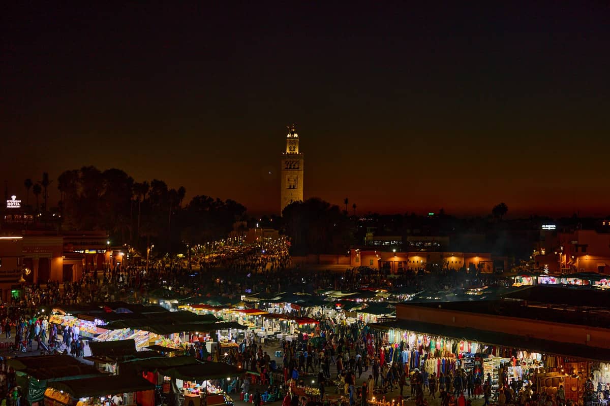 จัตุรัส Jemaa el-Fnaa กลางคืนที่มาร์ราเกช เต็มไปด้วยแผงขายของและผู้คน คูตูเบียมอสก์ส่องไฟอยู่ด้านหลัง