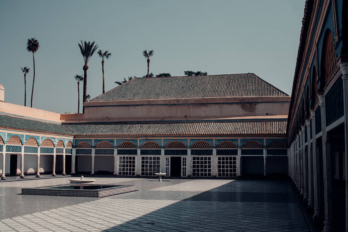 วิวกว้างของลานภายในพระราชวัง Bahia ในเมือง Marrakech ที่มีพื้นกระเบื้อง โค้งสีน้ำเงินและสีเหลือง และต้นปาล์มในพื้นหลัง