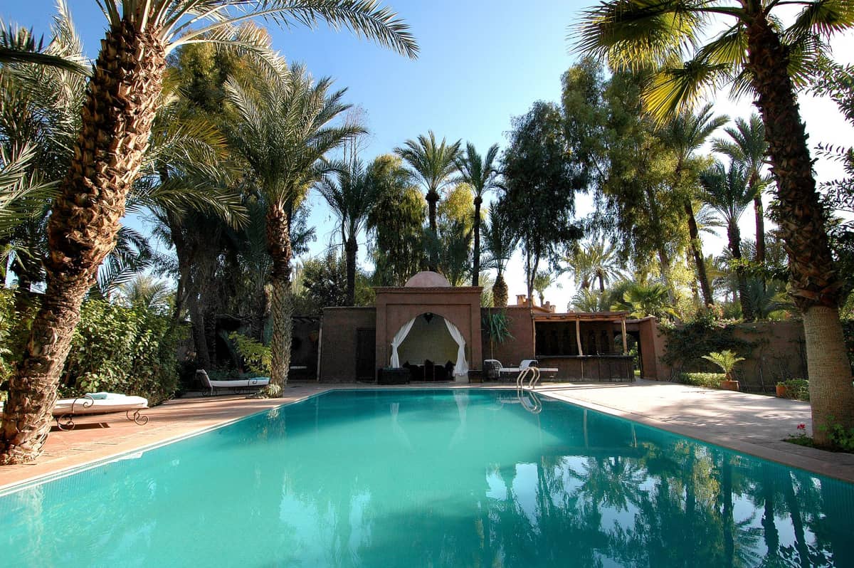 สระว่ายน้ำสีเทอร์ควอยซ์ขนาดใหญ่ล้อมรอบด้วยต้นปาล์มและวิลล่าหรูหราภายใต้ท้องฟ้าสีฟ้าสดใส สื่อถึงรีสอร์ทส่วนตัวใน Marrakech