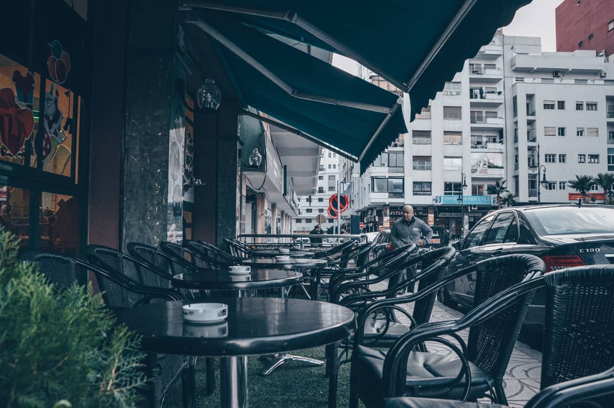 ที่นั่งกาแฟกลางแจ้งพร้อมโต๊ะและเก้าอี้สีดำบนถนนในเมืองที่เรียงรายด้วยอาคารสมัยใหม่ บรรยากาศชวนนึกถึงบาร์หรือเลานจ์ท้องถิ่น