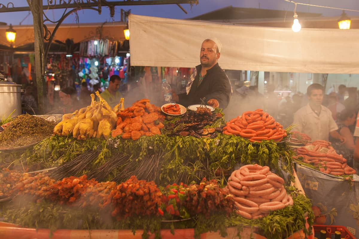 แผงอาหารในตลาดกลางคืนของ Marrakech ที่คึกคักพร้อมพ่อค้าและการจัดแสดงเนื้อสด ไส้กรอก สมุนไพร และผักขนาดใหญ่ที่ล้อมรอบด้วยไอน้ำ