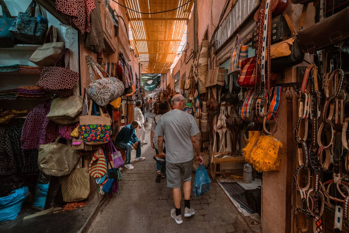 ซอยคึกคักในตลาดซูกของ Marrakech ที่เรียงรายด้วยกระเป๋าสีสันสดใส สินค้าหนัง และสิ่งทอ พร้อมนักช้อปที่กำลังเลือกดูสินค้าตามแผงต่างๆ