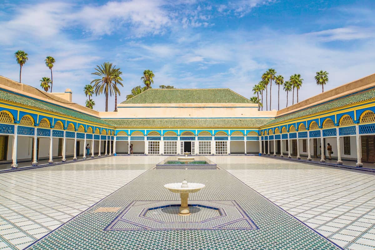 ลานกว้างปูกระเบื้องของพระราชวัง Bahia ในเมือง Marrakech พร้อมน้ำพุกลาง โค้งประตูสีฟ้าและเหลืองสวยงาม และต้นปาล์มใต้ท้องฟ้าสีคราม