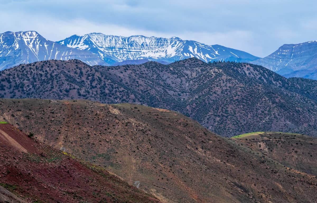 วิวภูเขา High Atlas ที่ปกคลุมด้วยหิมะพร้อมเชิงเขาที่ขรุขระเป็นสีเขียวและน้ำตาลภายใต้ท้องฟ้าที่มีเมฆมาก