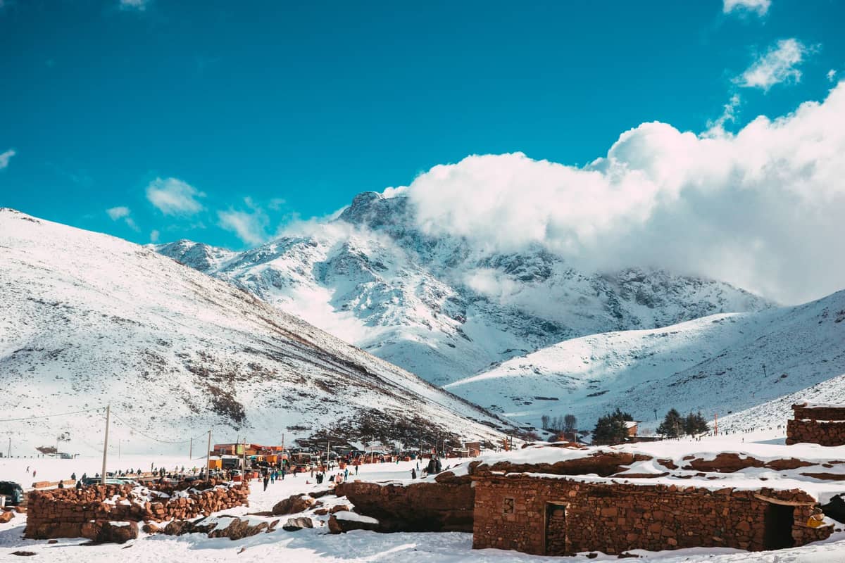ภูเขา High Atlas ที่ปกคลุมด้วยหิมะใต้ท้องฟ้าสดใส อาคารหินสีน้ำตาลในเบื้องหน้า มองเห็นนักเดินทางเขา; ความแตกต่างตามฤดูกาลที่ชัดเจนสื่อถึงการเปลี่ยนแปลงของสภาพอากาศ