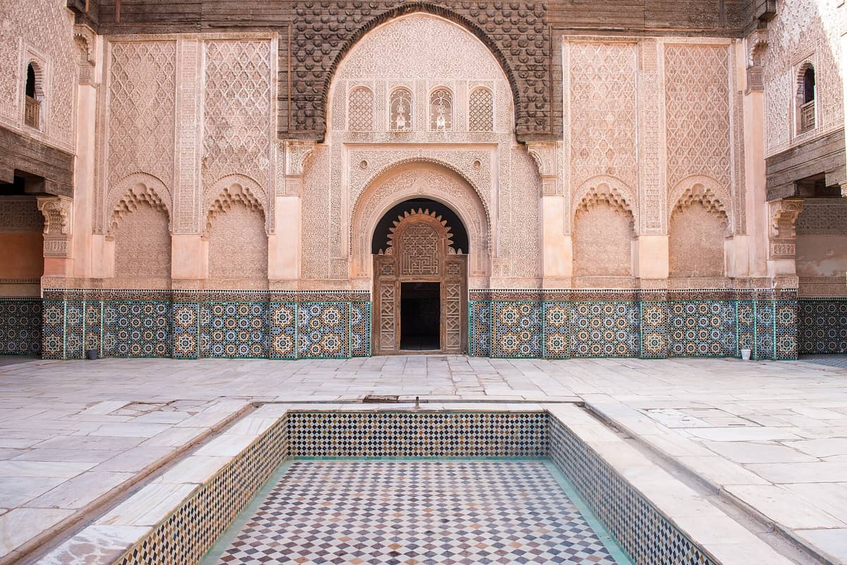 ลานภายในมาดราซา Ben Youssef ในเมือง Marrakech ที่ประดับด้วยปูนปั้นอันวิจิตร โค้งประตู งานกระเบื้องโมเสก และสระน้ำกลางที่ว่างเปล่า