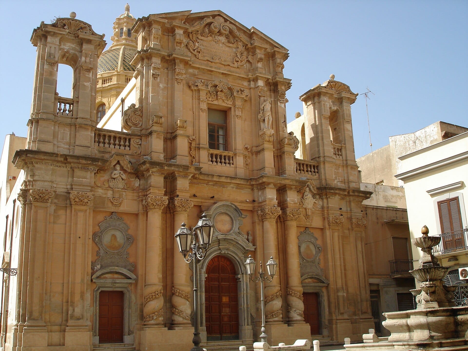 Barocke Fassade der Kathedrale von Marsala mit detaillierter Steinmetzarbeit, Bogentüren und einem Kuppelturm unter dem klaren blauen Himmel Siziliens.