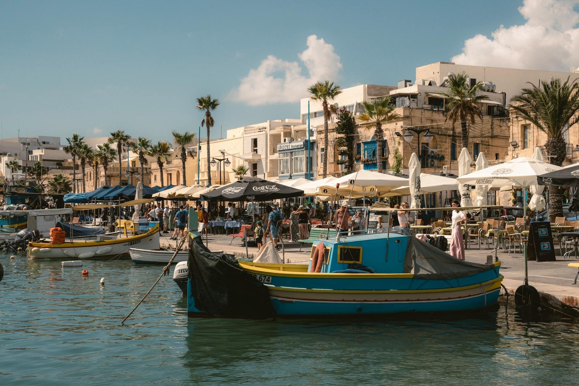 Tradizionali barche da pesca dai colori vivaci ormeggiate lungo il lungomare di Marsaxlokk, con banchi del mercato affollati, caffè e palme sullo sfondo.