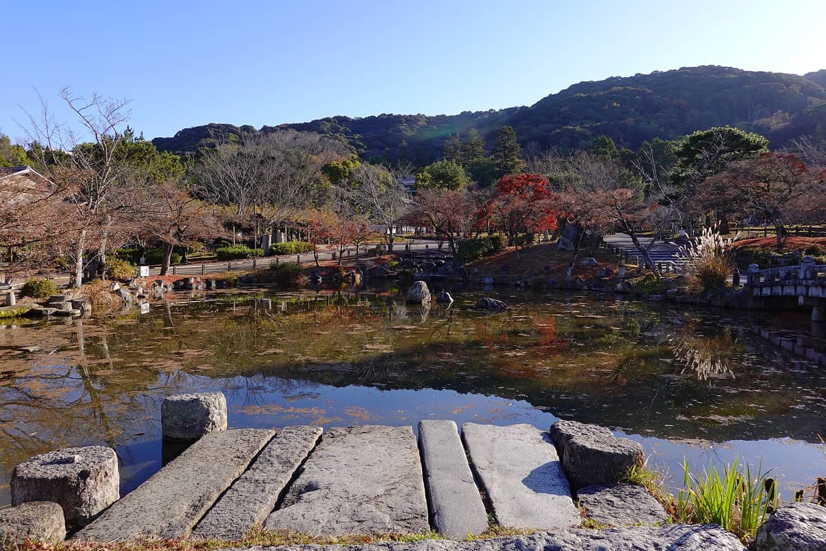 Scenic view of Maruyama Park pond framed by stone walkway, autumn trees, and distant forested hills under a bright blue sky in Kyoto.