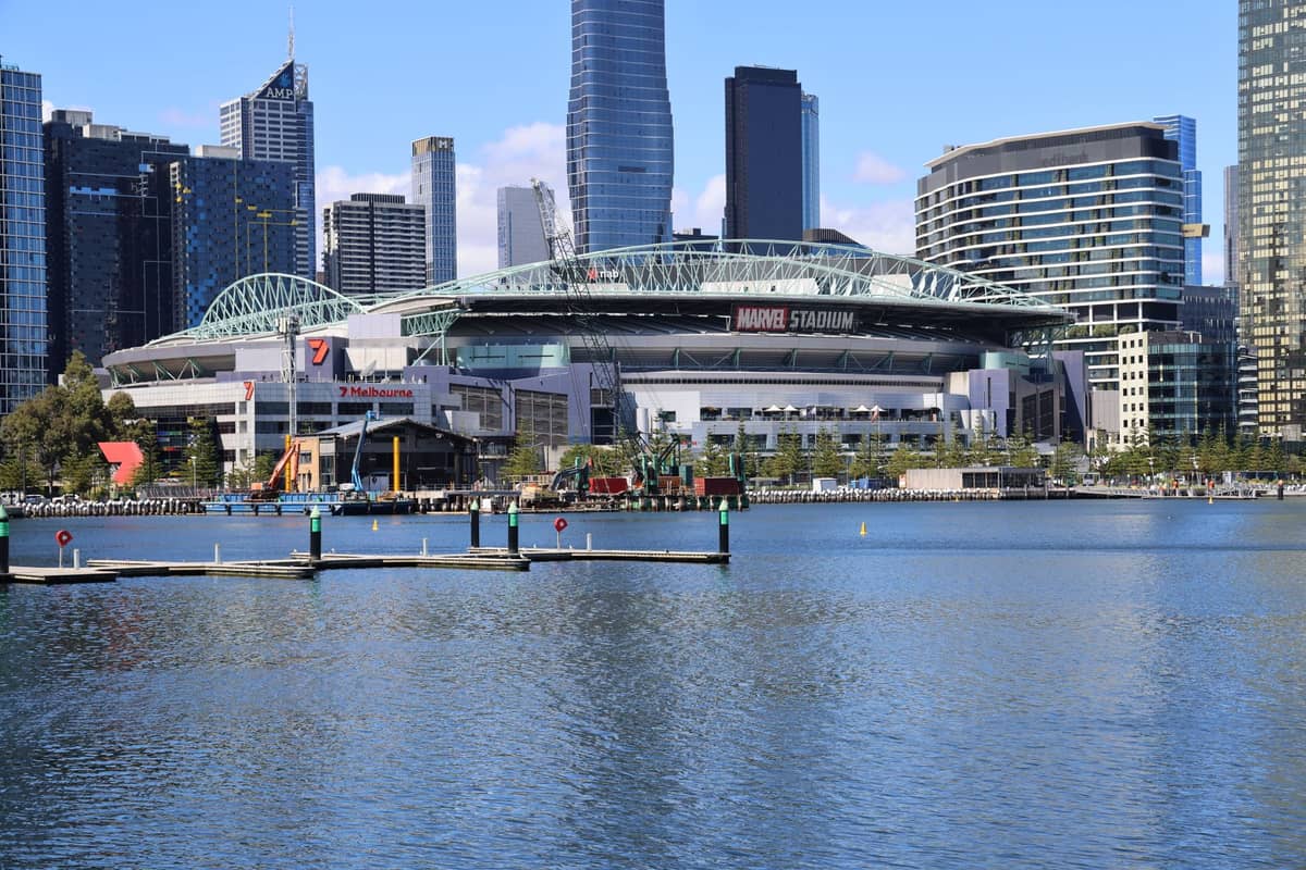 Das Marvel Stadium in Melbourne, vom Wasser aus fotografiert, mit der Skyline der Stadt und den Docklands-Gebäuden im Hintergrund an einem klaren Tag.