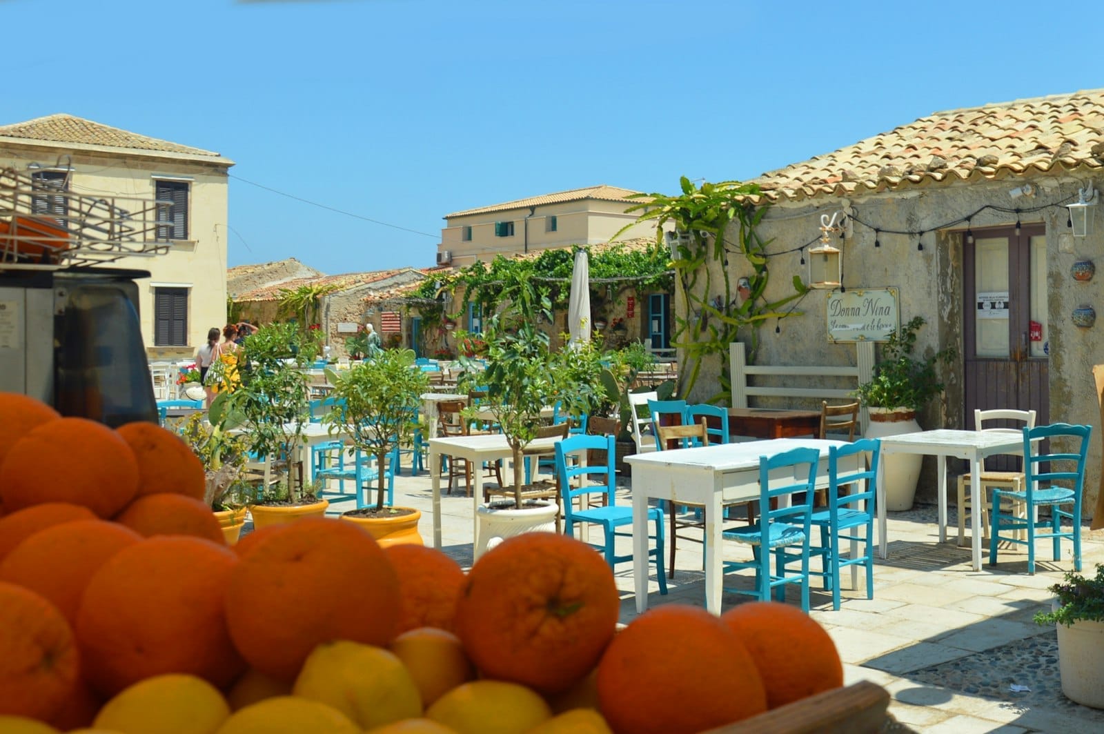 Outdoor-Café-Tische mit türkisfarbenen Stühlen und Topfpflanzen auf dem historischen Platz von Marzamemi, Zitrusfrüchte im Vordergrund, unter der hellen Mittagssonne.