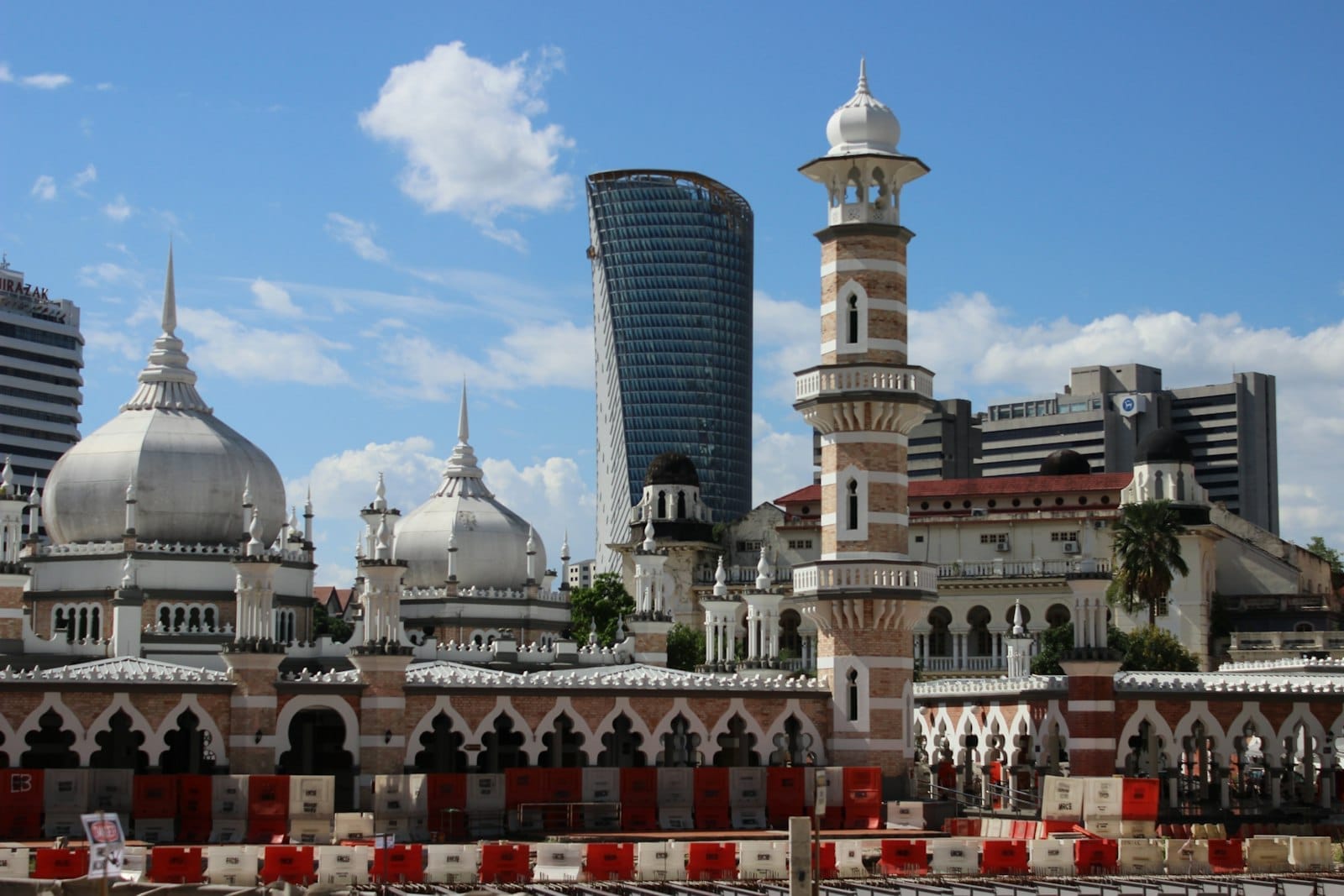 Masjid Jamek’s Mughal-style domes and tall minaret stand under a blue sky, with modern Kuala Lumpur skyscrapers in the background.