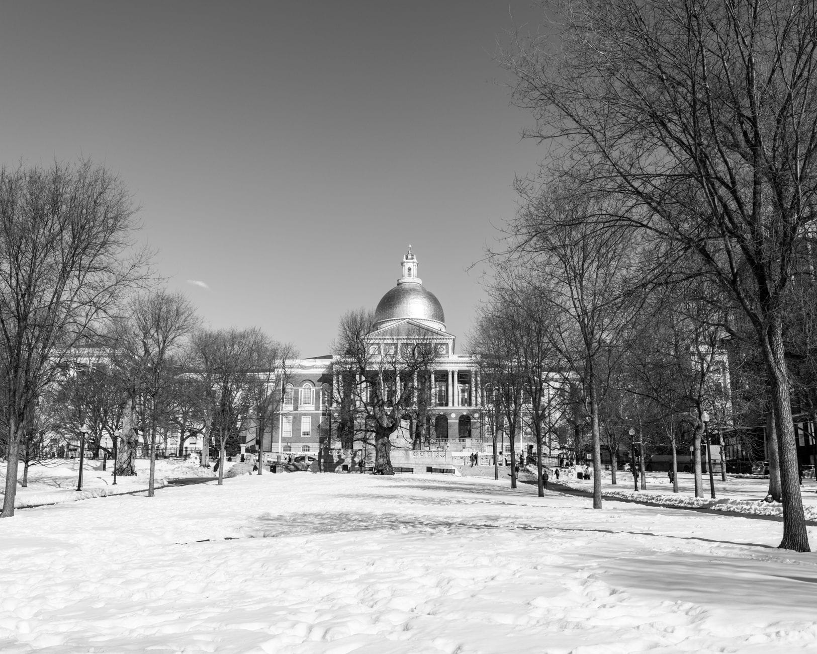 Scena invernale in bianco e nero del Massachusetts State House con la sua iconica cupola dorata, incorniciata da un Boston Common innevato e alberi spogli.
