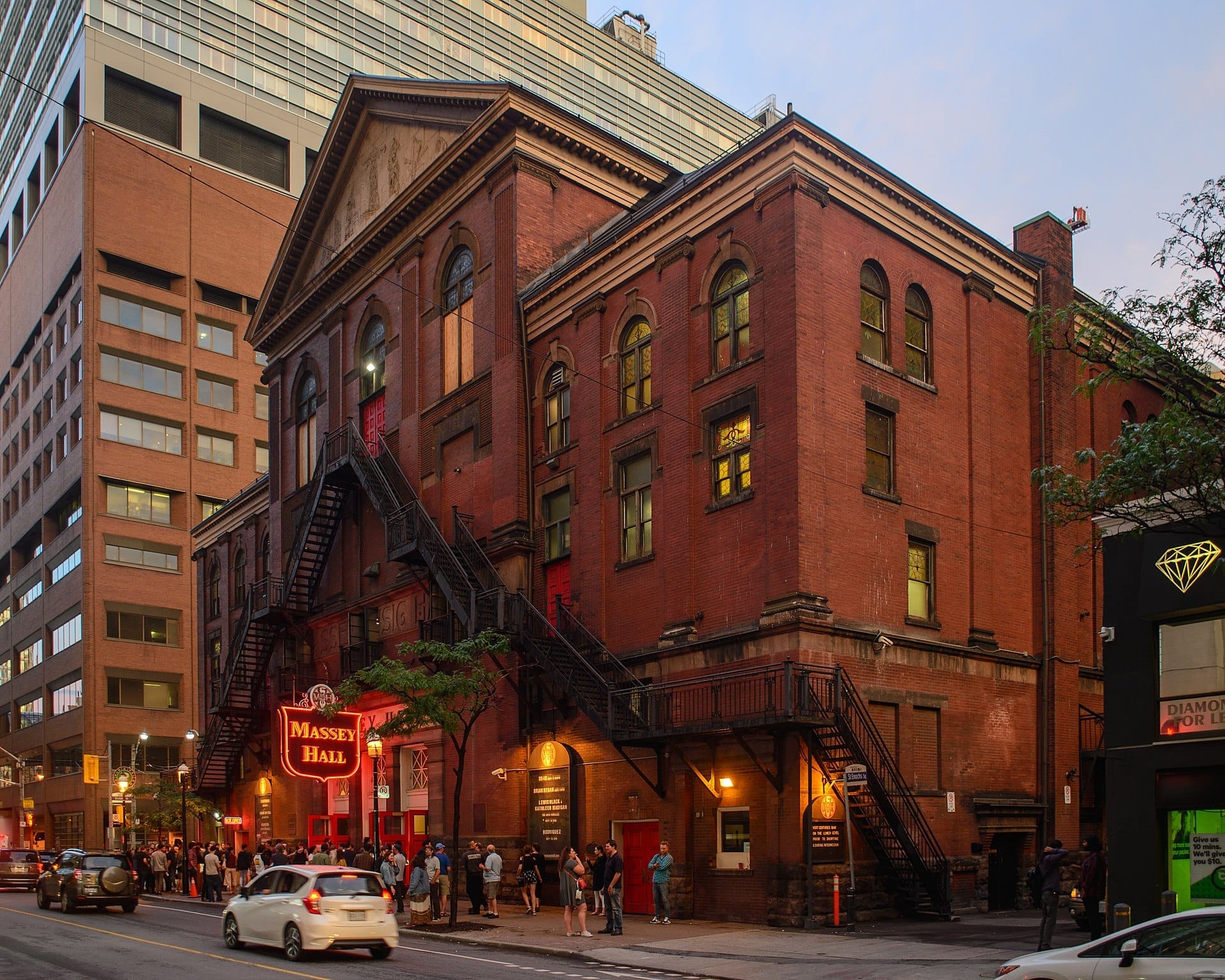 ด้านนอกของ Massey Hall ที่ทำจากอิฐแดงทางประวัติศาสตร์ในยามพลบค่ำ พร้อมป้ายนีออนและผู้คนที่เข้าคิวบนทางเท้าในใจกลางเมือง Toronto