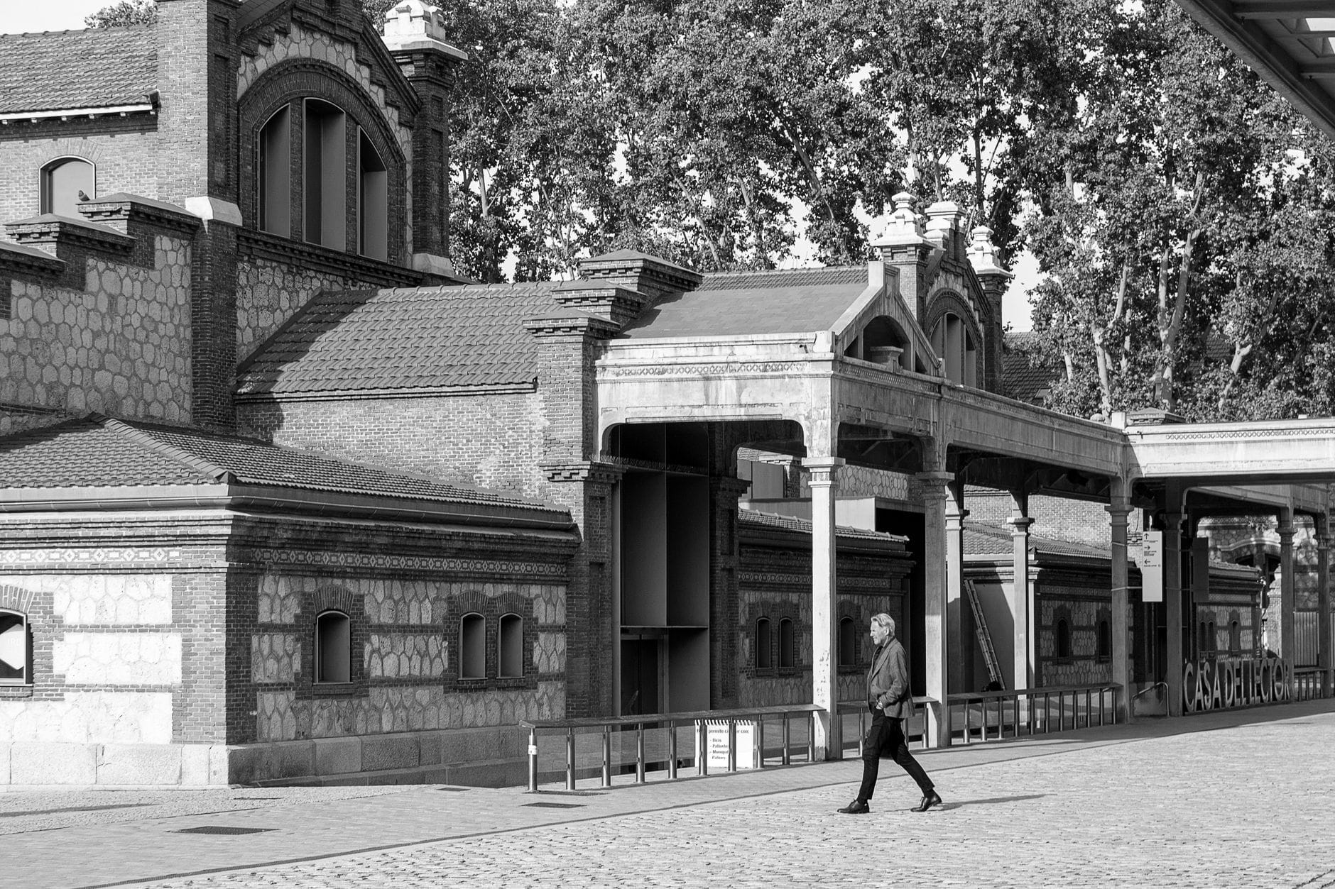 Foto in bianco e nero degli edifici originali di Matadero Madrid in stile neo-mudéjar, con una figura solitaria che cammina sulla piazza acciottolata.