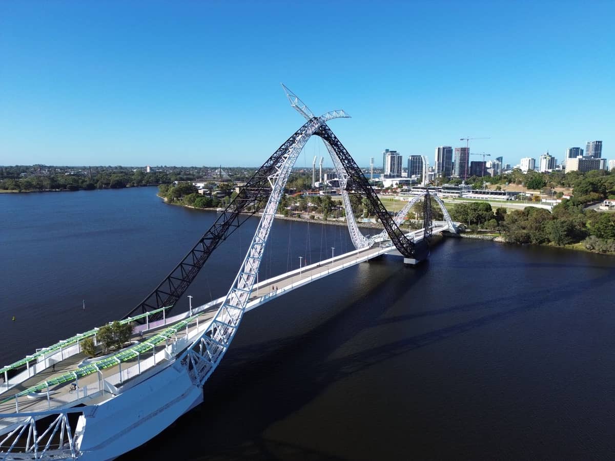 Luftaufnahme der Matagarup Bridge in Perth mit ihrem markanten Bogenbauwerk über dem Swan River – im Hintergrund die Stadtsilhouette unter strahlend blauem Himmel.