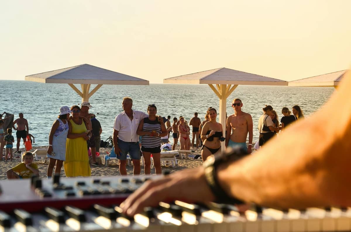 Nahaufnahme eines Keyboardspielers an einem belebten Strand mit versammelten Menschen, Sonnenschirmen und dem Meer im Hintergrund bei Tageslicht.