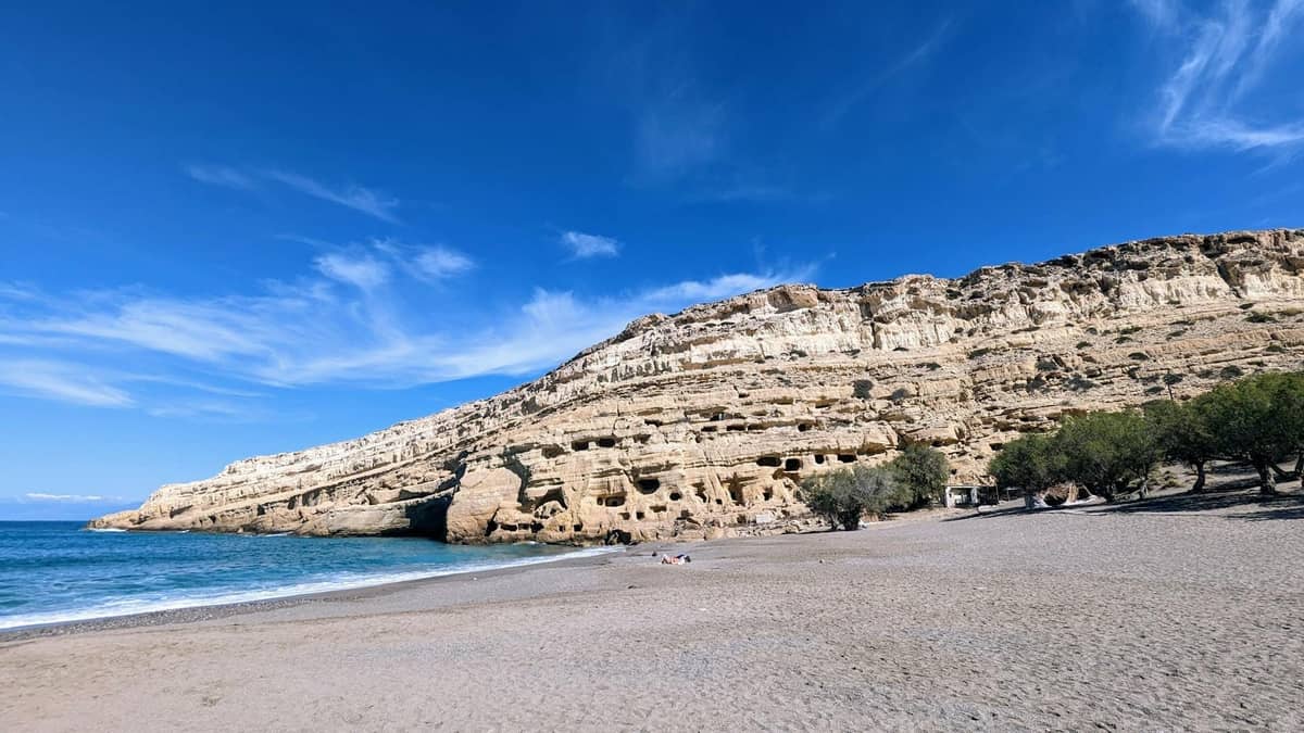 Weitwinkelansicht des Strandes Matala mit den markanten Sandsteinfelswänden und sichtbaren Höhlen im Fels unter blauem Himmel.