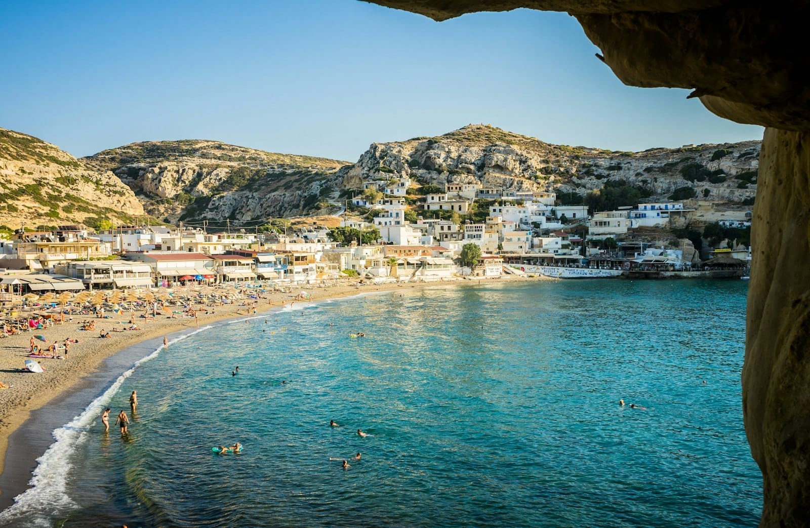 La spiaggia di Matala con acqua turchese, bagnanti, sabbia dorata, ombrelloni e le famose grotte nella falesia, vista dall'interno di una grotta in una luminosa giornata di sole.