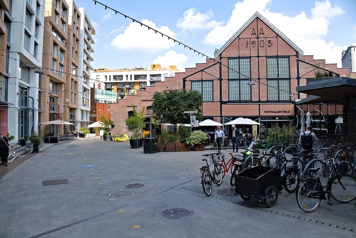 ด้านหน้า Mathallen Oslo ฟู้ดฮอลล์อิฐเก่าติดป้ายปี '1908' มีผู้คนเดินเล่น ปั่นจักรยาน ที่นั่งกลางแจ้ง อาคารสมัยใหม่ในเขต Vulkan ที่ปรับปรุงใหม่ล้อมรอบ