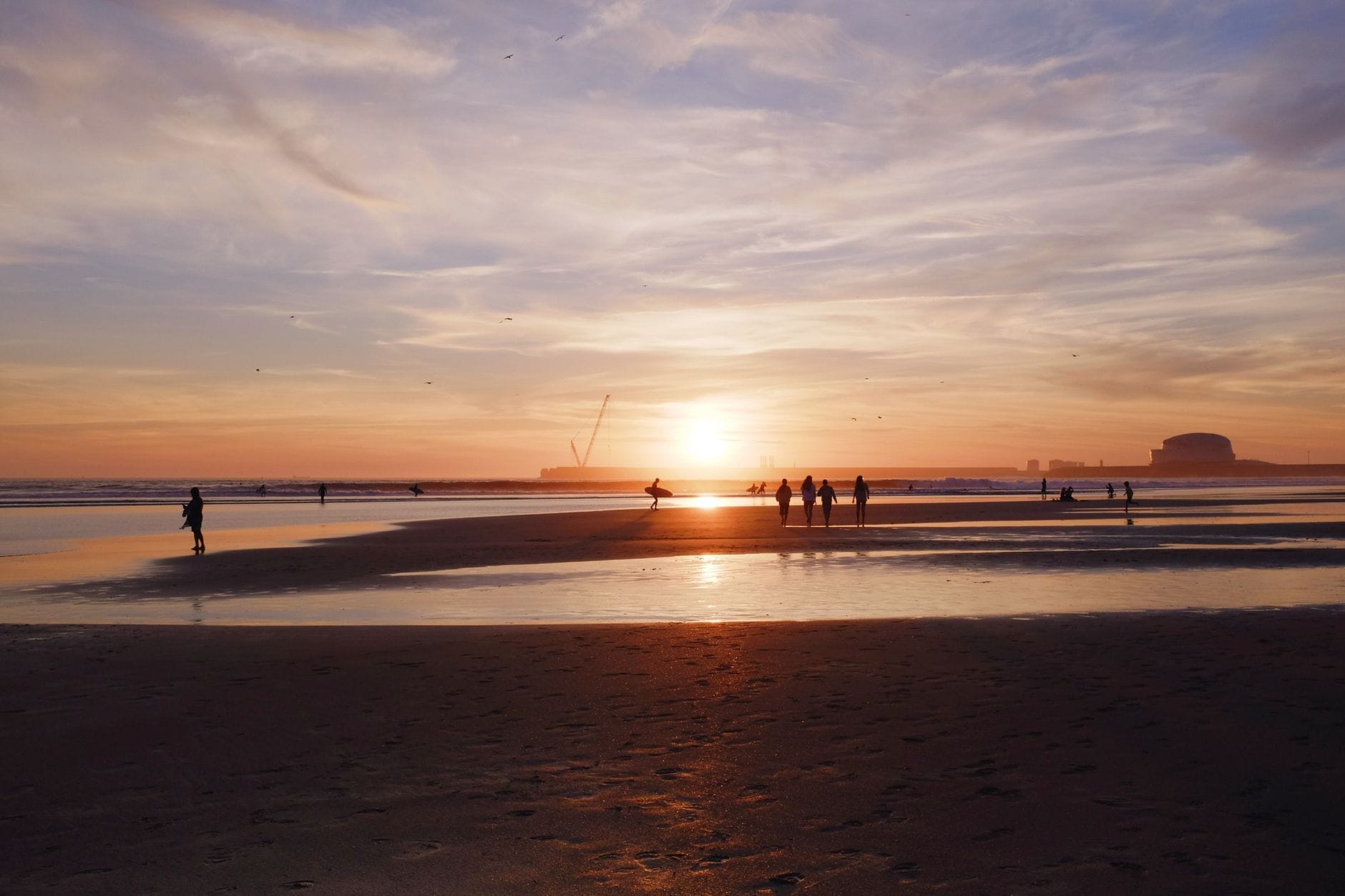 Gruppi di persone che godono di un tramonto dorato sull'ampia distesa sabbiosa della spiaggia di Matosinhos, con le onde dell'Atlantico e le strutture portuali sullo sfondo.