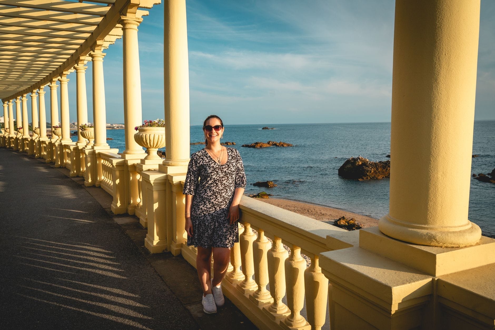 Donna in piedi sul lungomare di Matosinhos, incorniciata da colonne gialle, con vista sulla Praia de Matosinhos e la costa rocciosa sullo sfondo.