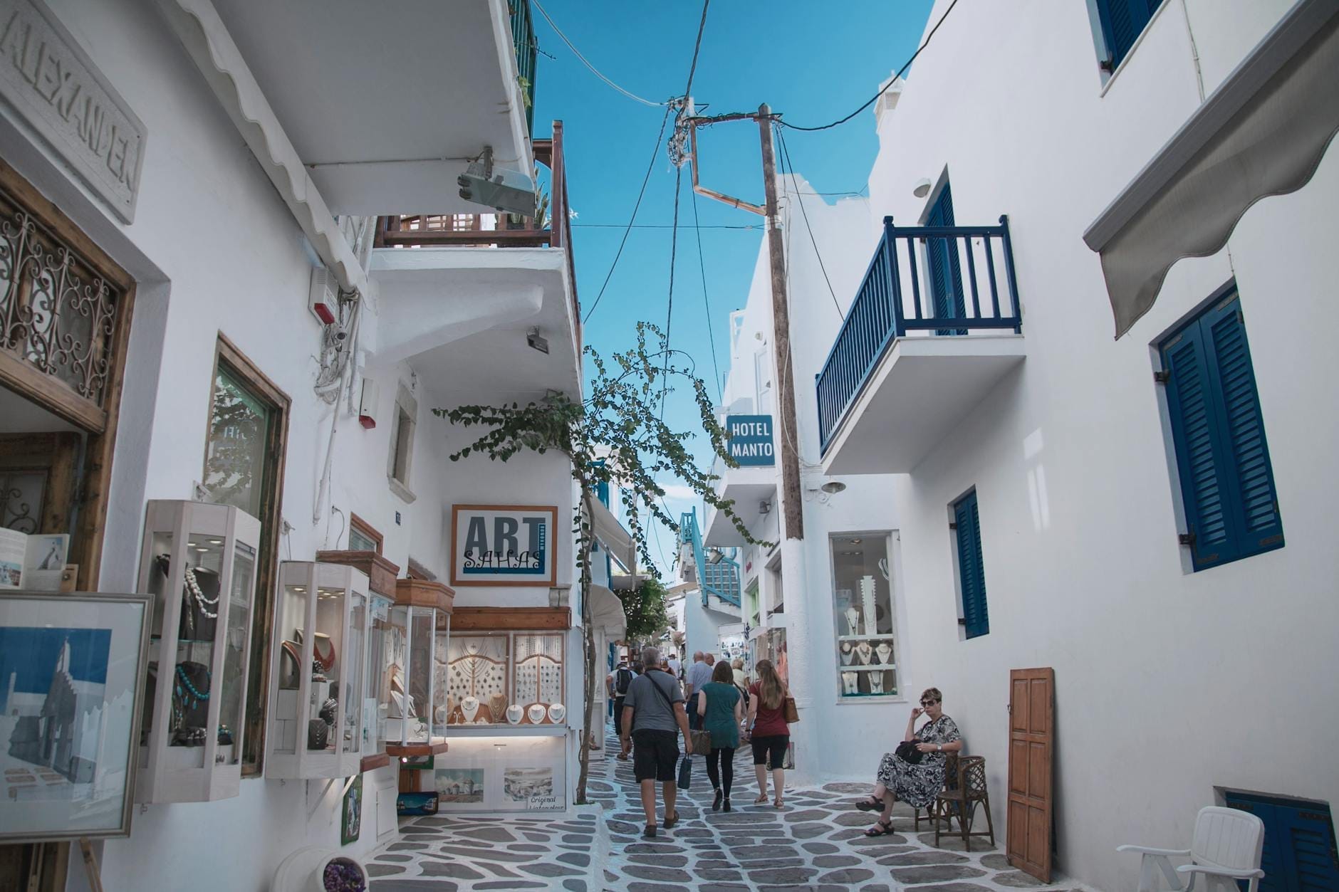 Personas paseando por la calle Matoyianni en Mykonos Town, rodeadas de edificios cicládicos blancos, boutiques y cafés bajo un cielo azul despejado.