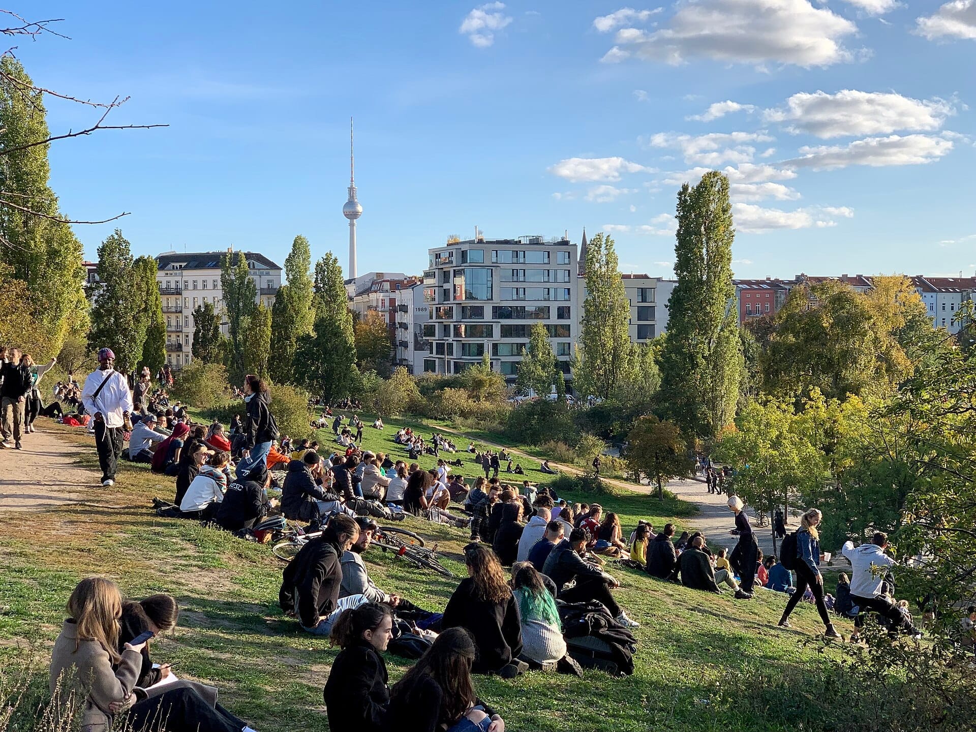 Folle di persone che si rilassano e socializzano sui prati in pendenza del Mauerpark di Berlino, con gli edifici della città e la torre televisiva visibili sotto un cielo blu.