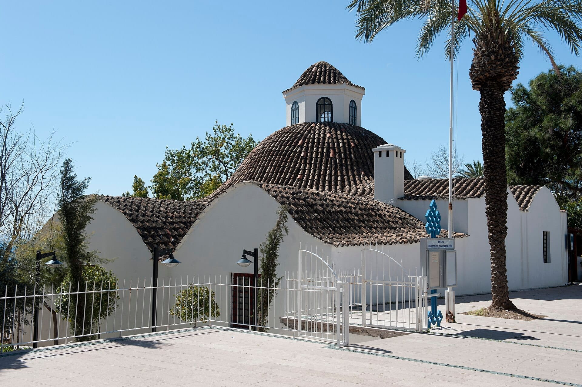 Il Museo del Convento Mevlevi di Antalya presenta mura imbiancate e un tradizionale tetto a cupola, incorniciato da palme e immerso nella luce del giorno.