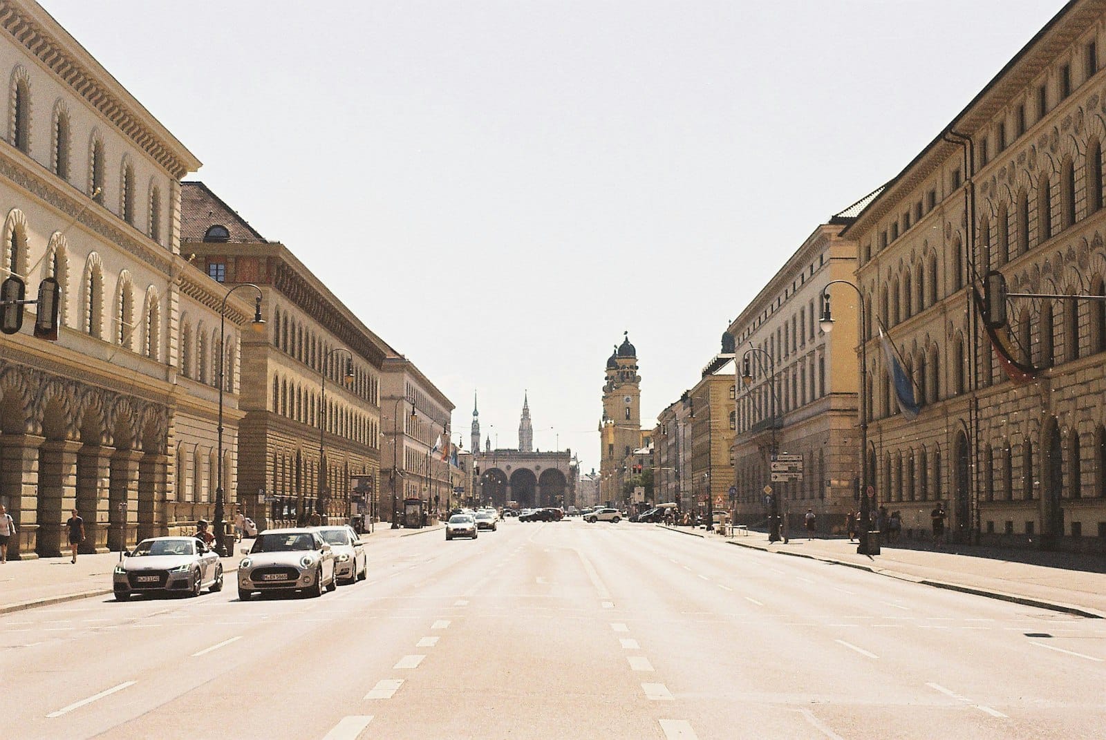 มุมมองกว้างตามถนน Ludwigstrasse ใน Maxvorstadt, Munich เห็นอาคารนีโอคลาสสิก รถยนต์ ซิเกสทอร์และโบสถ์ Theatiner อยู่ไกล ๆ ในวันที่มีแดด