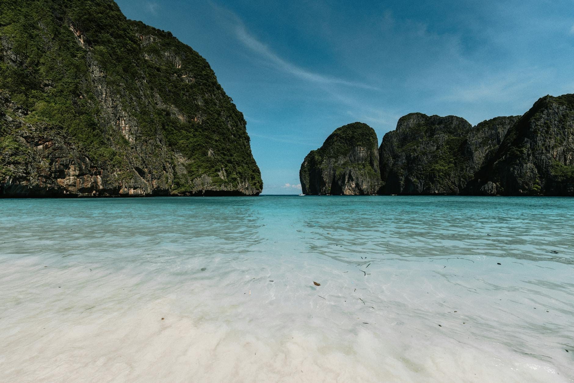 Wide view of Maya Bay’s turquoise water and white sandy beach framed by dramatic limestone cliffs under a clear blue sky, with no people visible.