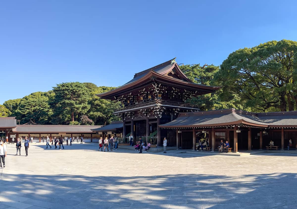 Wide view of Meiji Jingu Shrine’s main wooden gate and courtyard surrounded by trees under a clear blue sky with visitors walking around.