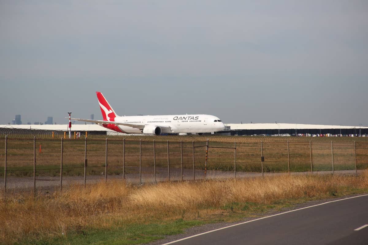 Ein Qantas-Flugzeug auf der Startbahn mit der Skyline von Melbourne, die in der Ferne schwach zu sehen ist, aufgenommen hinter einem Flughafensicherheitszaun.