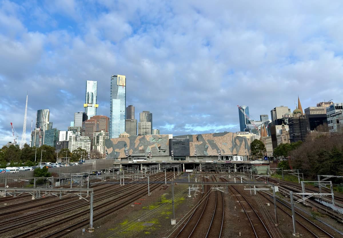 Ampia veduta dello skyline di Melbourne con moderni grattacieli e la caratteristica facciata di Federation Square oltre i binari ferroviari sotto un cielo prevalentemente nuvoloso.