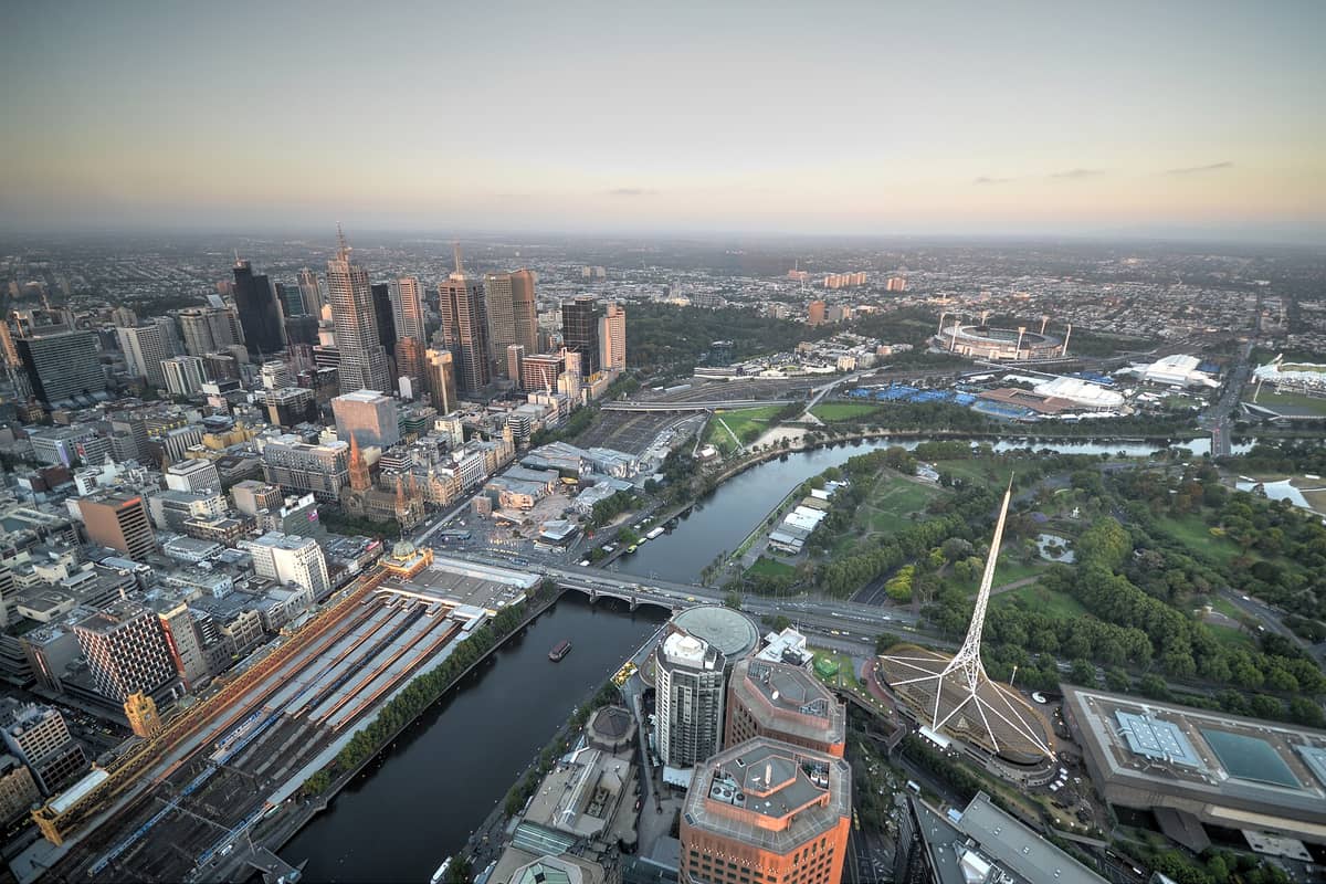 ภาพมุมสูงของย่าน Melbourne CBD ยามพระอาทิตย์ตก เห็นผัง Hoddle Grid แม่น้ำยาร์รา สถานีรถไฟ Flinders Street ตึกสูงและสวนสาธารณะล้อมรอบ
