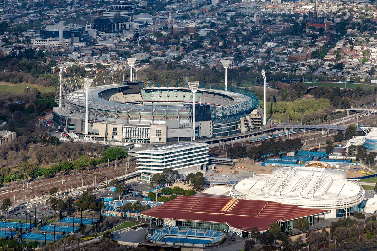 Vista aérea del Melbourne Cricket Ground (MCG) mostrando el estadio ovalado completo rodeado de parques, vías de tren y calles de la ciudad en un día despejado.