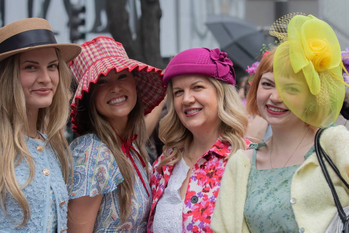 Vier Frauen in bunten Kleidern und festlichen Hüten, die bei einer Outdoor-Veranstaltung mit Stadtkulisse lächeln.