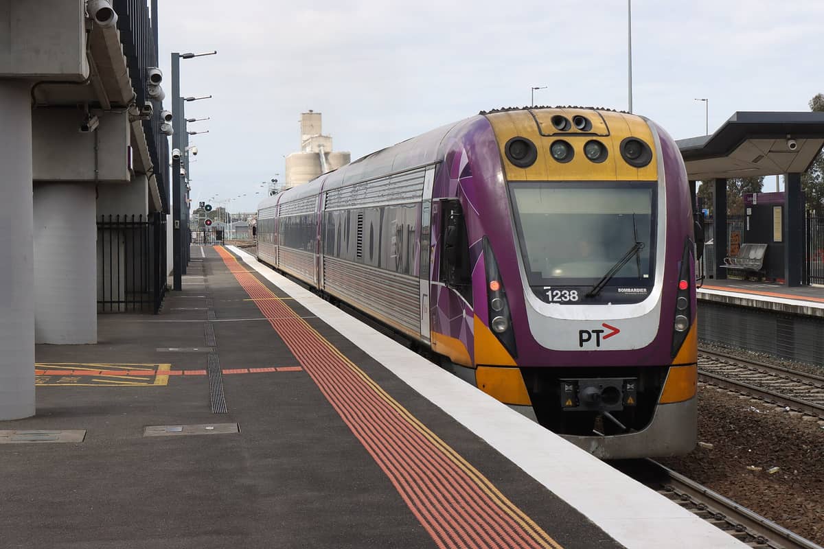 Ein V/Line-Zug am Bahnsteig des Bahnhofs Flemington Racecourse, mit sichtbaren Gleisen und Bahnhofseinrichtungen unter bewölktem Himmel.
