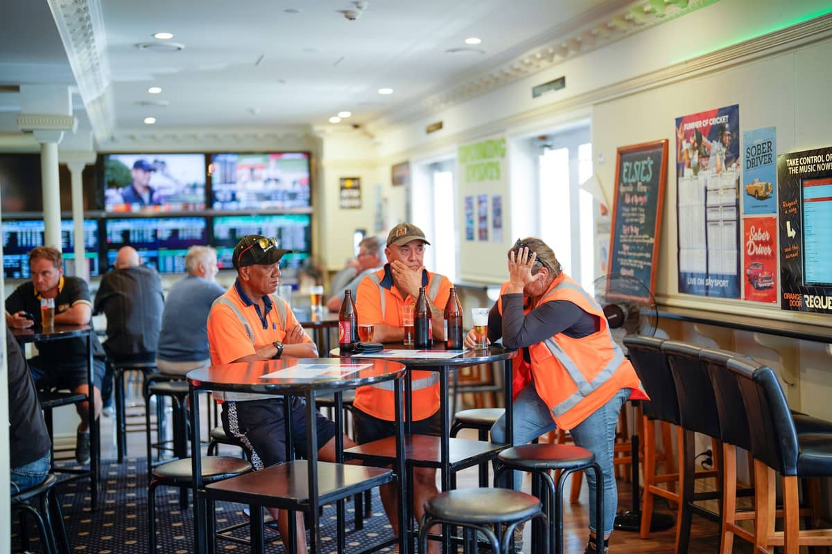 Personen in Warnwesten sitzen an Tischen in einem lebhaften Pub in Melbourne mit mehreren Fernsehbildschirmen im Hintergrund.