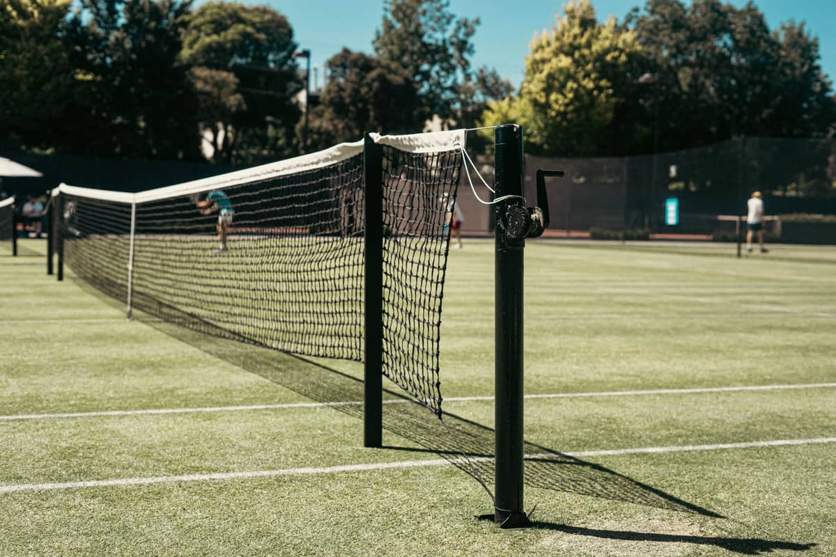 Tampilan jarak dekat lapangan tenis luar ruangan dengan orang-orang bermain di hari yang cerah, dikelilingi pepohonan.