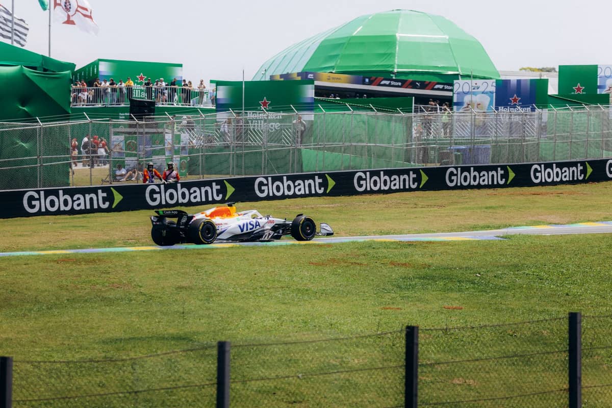Mobil balap Formula 1 di bagian berumput sirkuit dengan spanduk Globant dan Heineken yang terlihat serta penonton di struktur acara berwarna hijau.