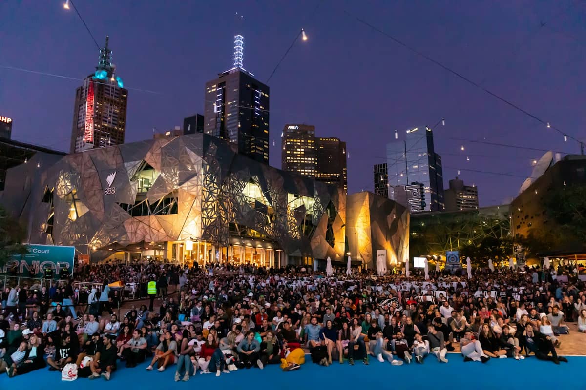 Kerumunan besar berkumpul di luar ruangan di Federation Square di pusat kota Melbourne saat senja, dengan gedung-gedung kota modern di latar belakang.