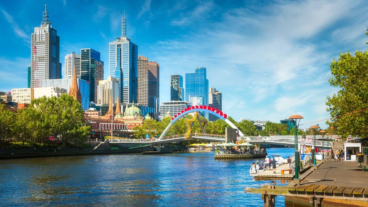 View of Melbourne skyline with the Yarra River in the foreground, arching bridge, riverside walk, and green trees on a bright sunny day.